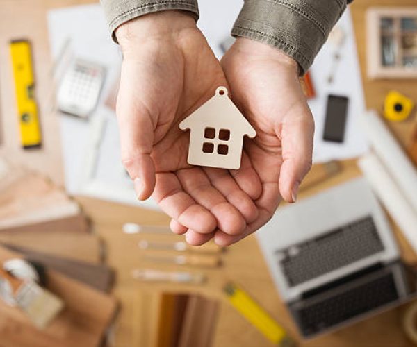 Real estate agent holding a small house, desktop with tools, wood swatches and computer on background, top view